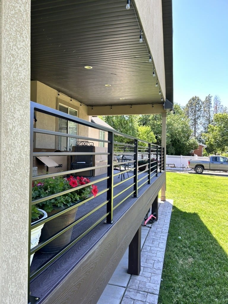 Black horizontal bar railing running along a covered deck with potted geraniums and a lawn beyond