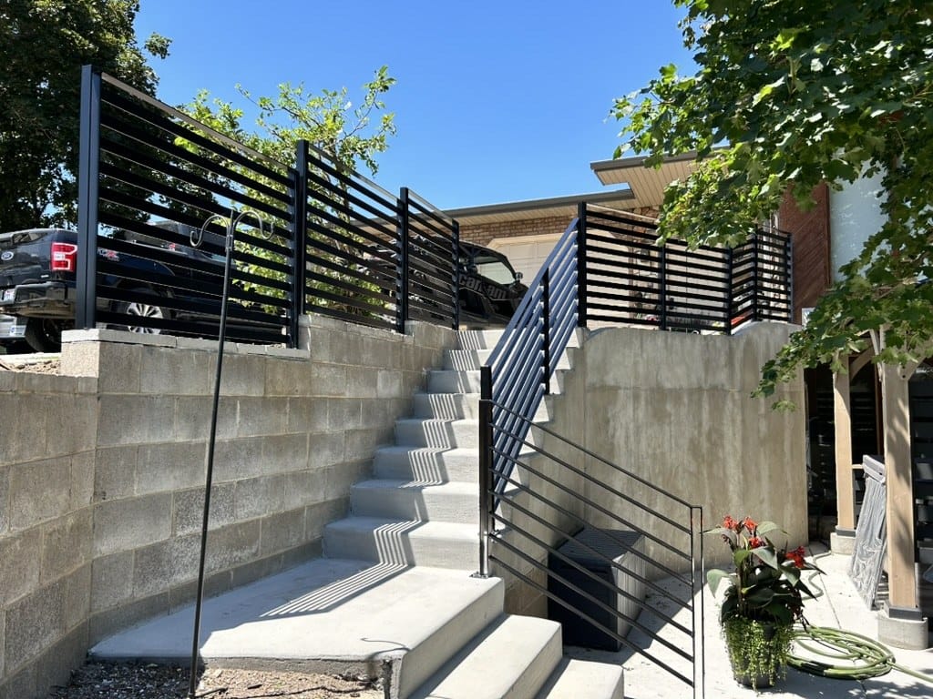 Black horizontal slat railing and privacy screen along an exterior stair run above a block retaining wall