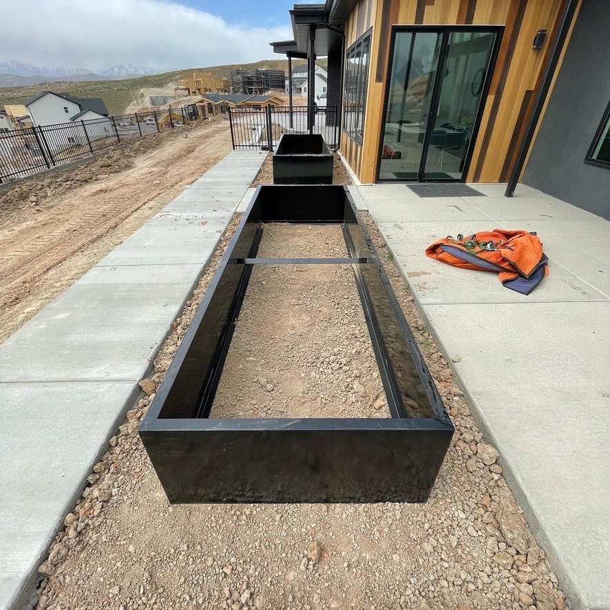Long black steel planter box with welded cross-bracing set in gravel on a Utah residential job site