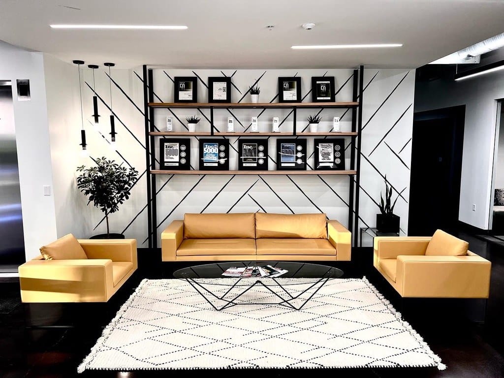 Black steel shelving frame with walnut shelves on a graphic accent wall above tan leather sofas in a commercial lobby