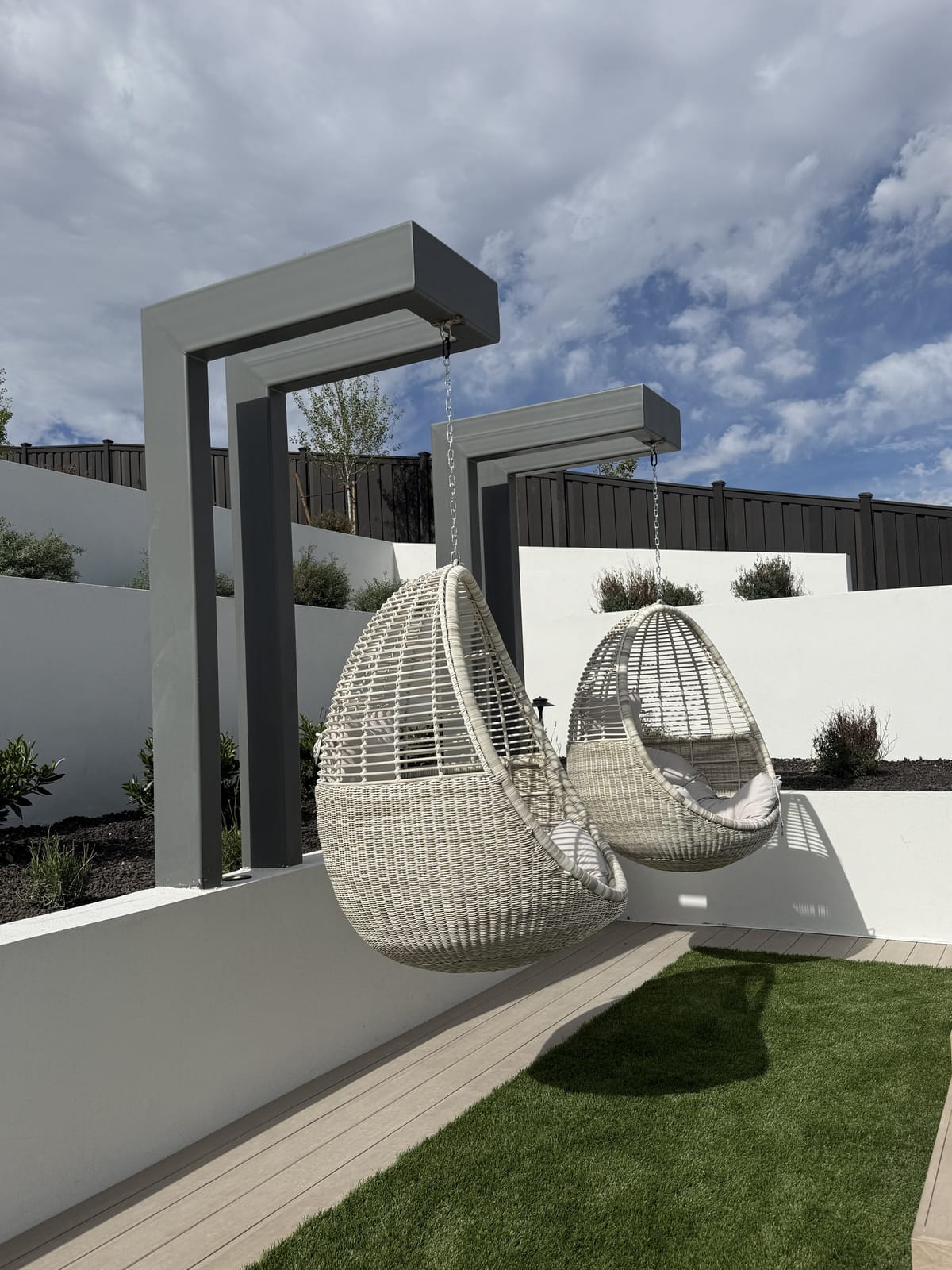 Two woven hanging egg chairs suspended from a grey powder-coated steel swing structure in a landscaped backyard
