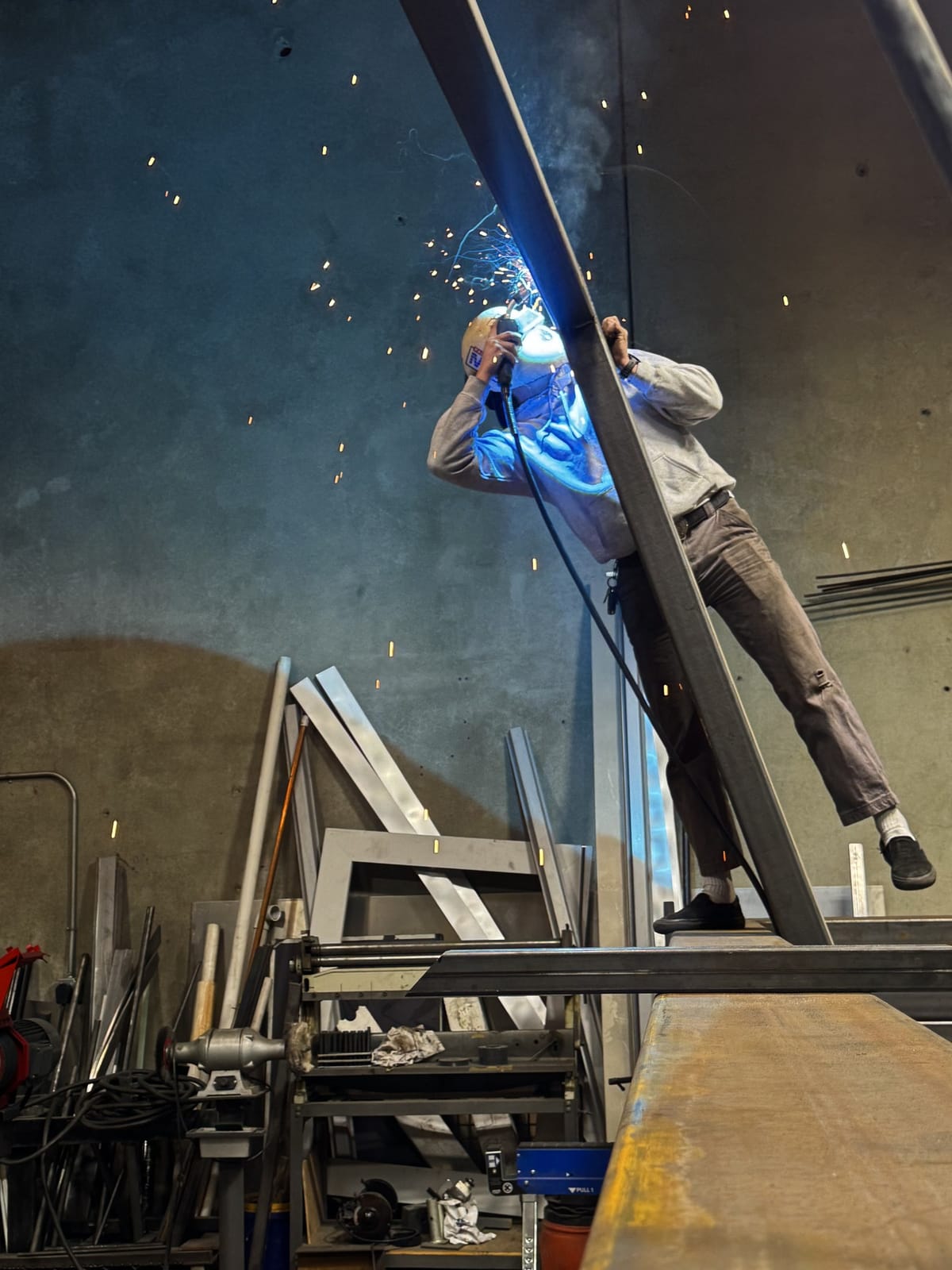 Denver welding a steel beam overhead in the Lehi shop, arc light and sparks falling against the concrete wall